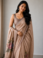 A woman modeling a draped beige saree with a matching sleeveless blouse. The saree has a subtle sheen and features floral embroidery in pink, green, and blue on the pallu end, set against a neutral indoor background.