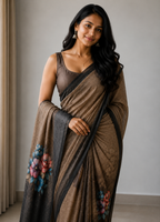 A woman modeling a brown and black saree with colorful floral embroidery near the border, paired with a matching brown blouse, in an indoor setting.