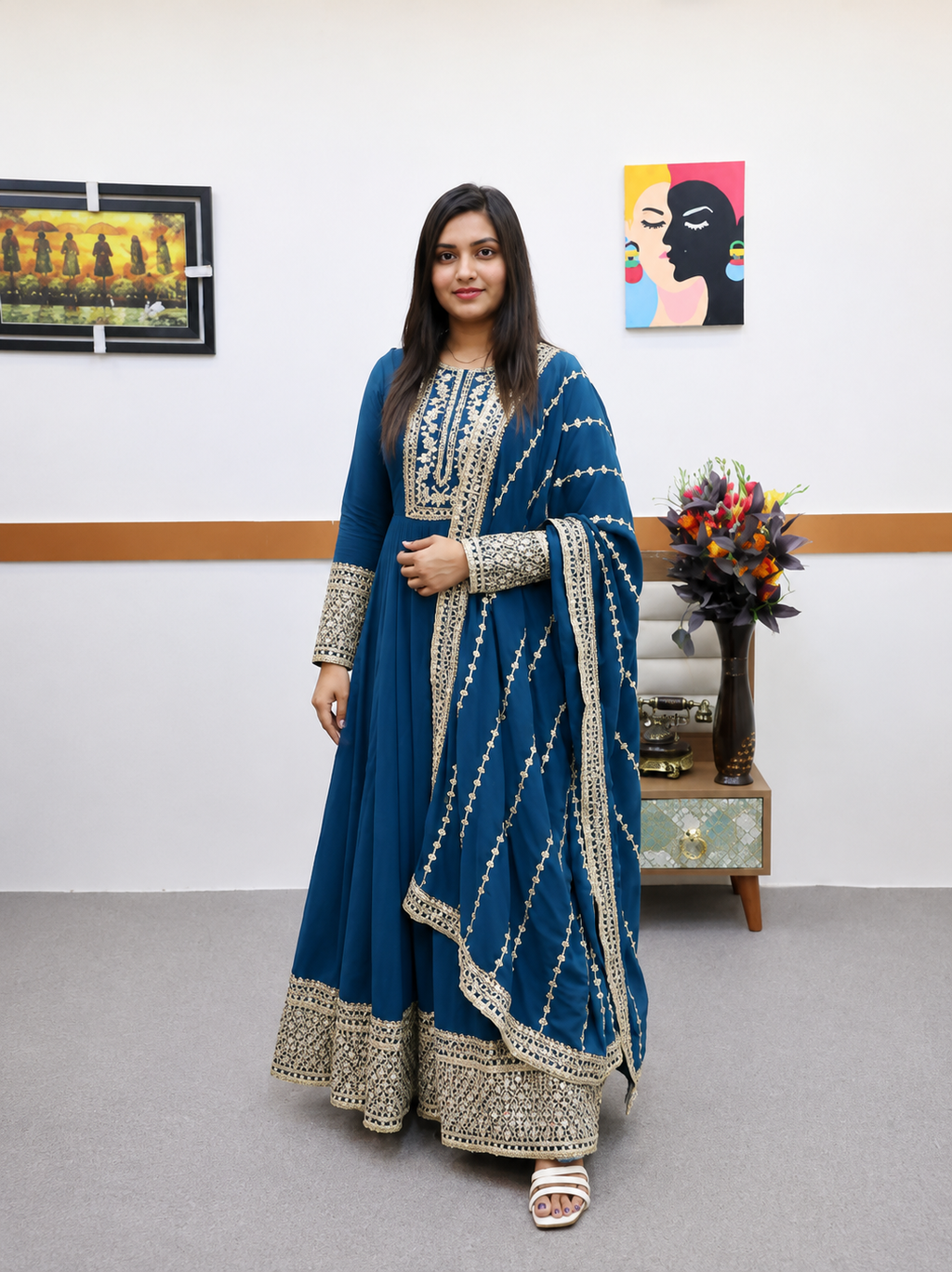 A woman modeling a traditional Indian outfit consisting of a teal blue anarkali kurta with intricate silver embroidery, paired with a matching dupatta, in an indoor setting.