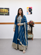 A woman modeling a traditional Indian outfit consisting of a teal blue anarkali kurta with intricate silver embroidery, paired with a matching dupatta, in an indoor setting.