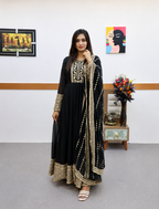 A woman modeling a black traditional Anarkali gown with extensive gold embroidery, paired with a matching embroidered dupatta, in an indoor setting with framed wall art and a small side table.