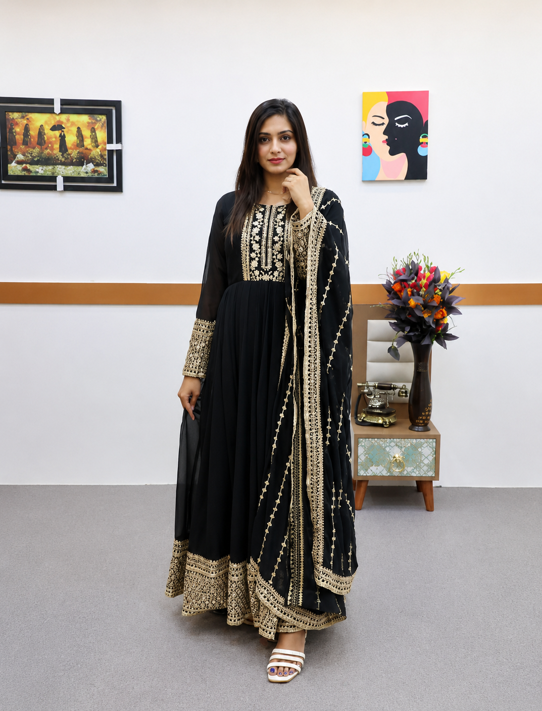 A woman modeling a black traditional Anarkali gown with extensive gold embroidery, paired with a matching embroidered dupatta, in an indoor setting with framed wall art and a small side table.