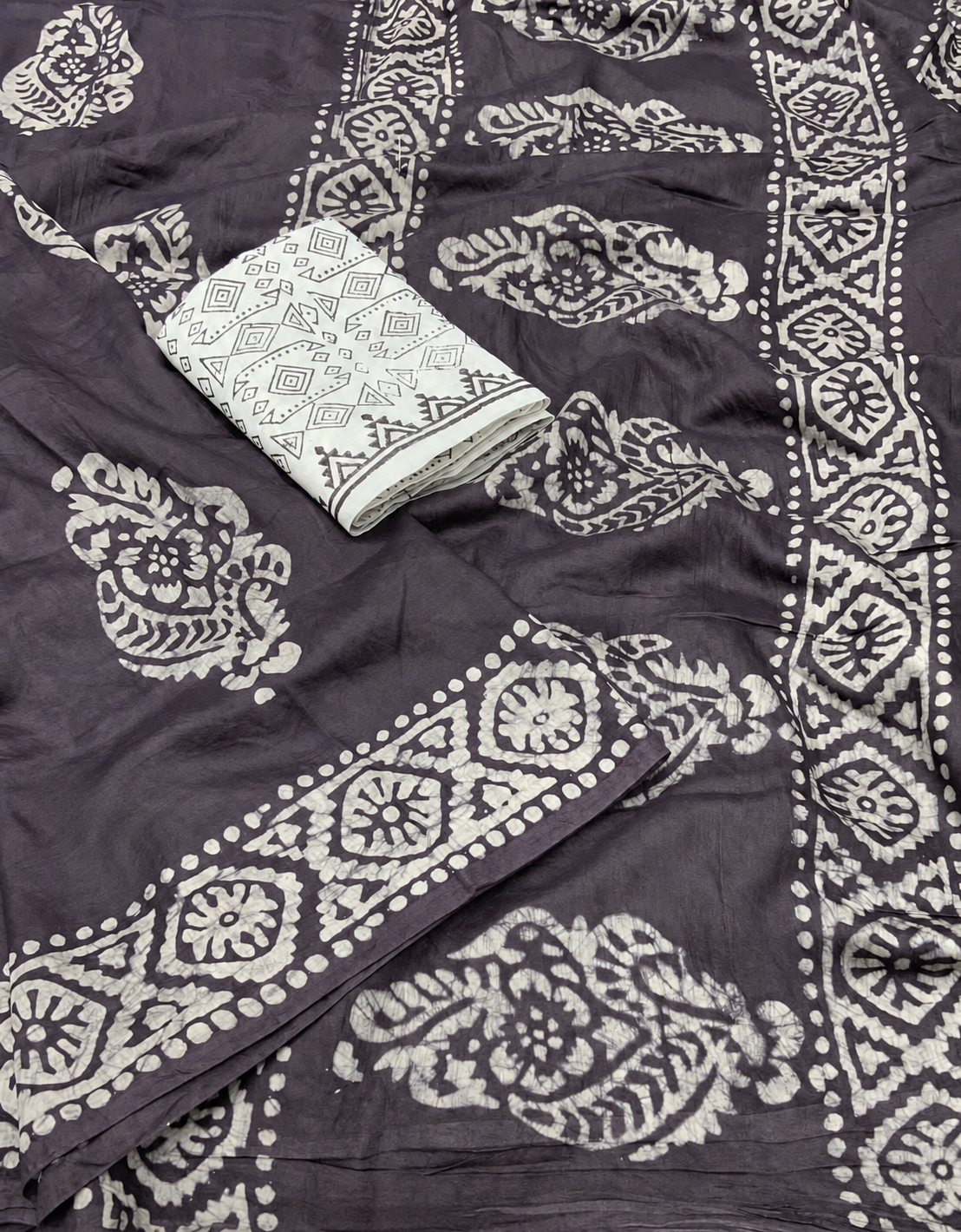 A folded and unfolded view of a dark gray block-printed fabric, likely cotton or silk, featuring traditional floral motifs and geometric borders in a contrasting off-white color.