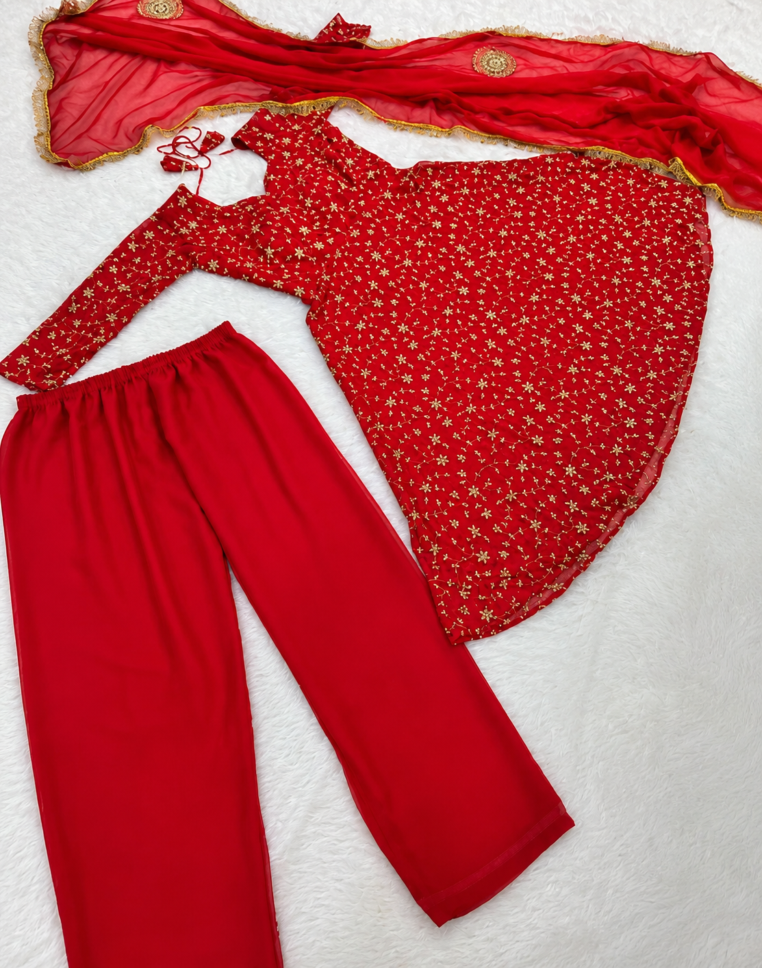 A red three-piece traditional outfit laid flat on a white textured background, including a long-sleeve top with a small gold floral print, solid red pants, and a matching sheer red dupatta with gold border detailing.