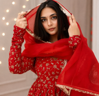 A woman wearing a traditional red embroidered dress with a matching sheer red dupatta and gold-trimmed veil, holding the veil in front of her face against a softly lit indoor background.