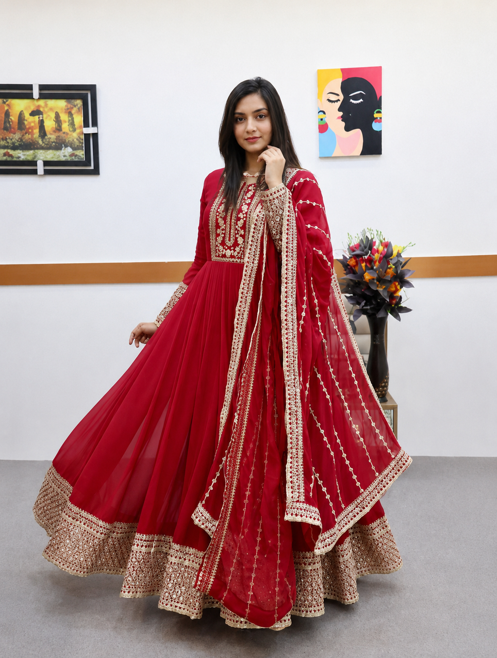 A woman modeling a traditional red anarkali gown with gold embroidery and a matching dupatta in an indoor studio setting.