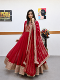 A woman modeling a traditional red anarkali gown with gold embroidery and a matching dupatta in an indoor studio setting.