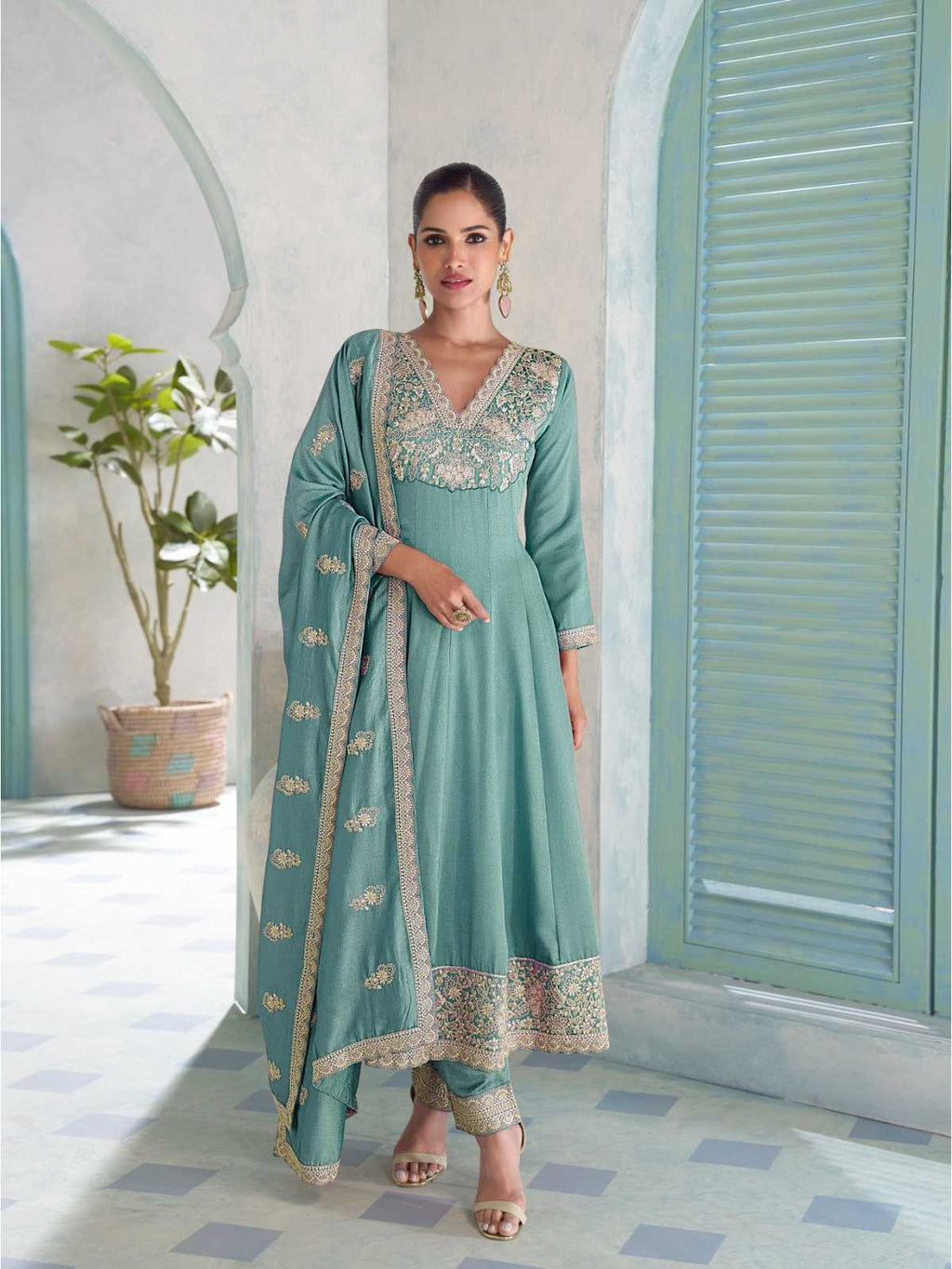 A woman models a mint green traditional Indian salwar kameez suit with intricate gold embroidery, standing in a courtyard with arches and shutters.