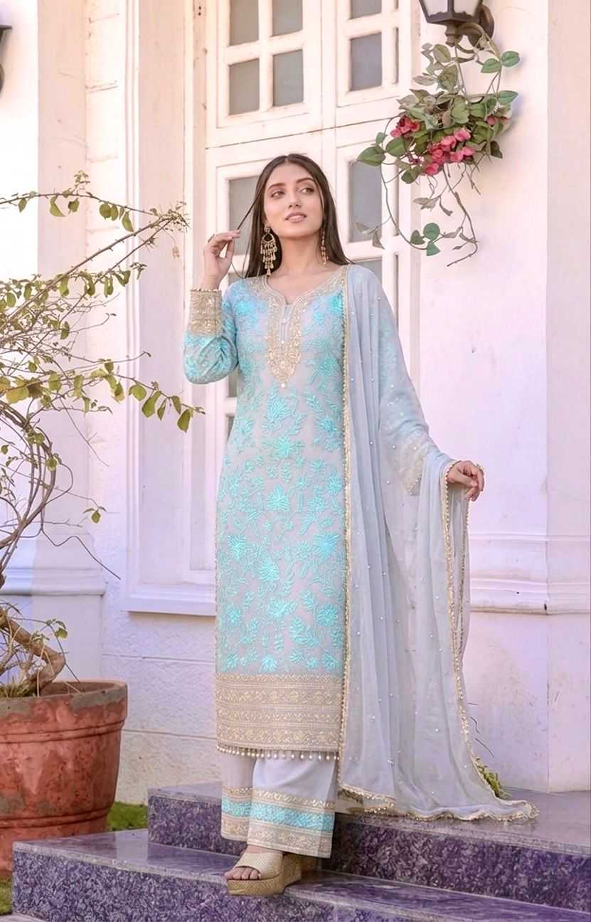 A woman models a traditional three-piece salwar kameez suit in light blue and white with intricate gold embroidery, standing on stone steps in front of a white building.