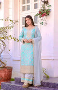 A woman stands on stone steps in front of a white building, wearing a light aqua blue traditional outfit with gold embroidery and a sheer light gray dupatta.