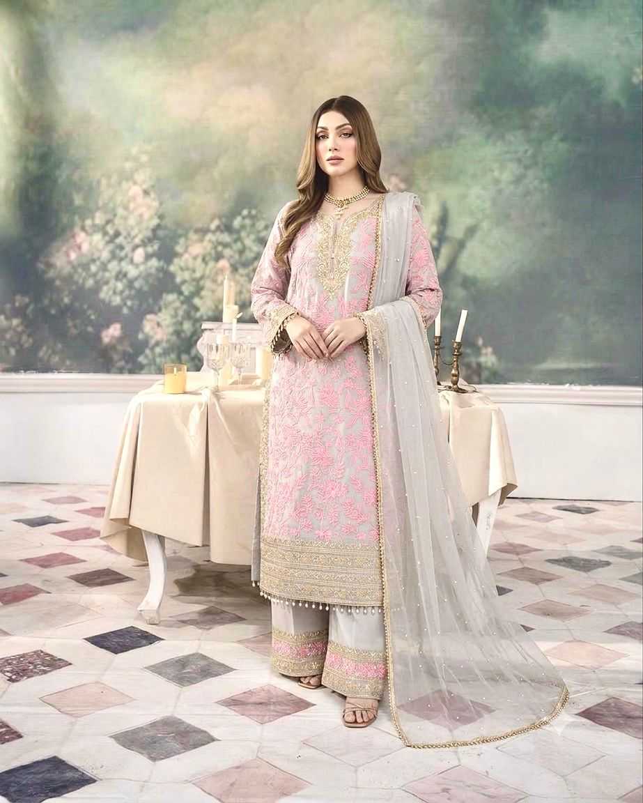 A woman models a traditional three-piece salwar kameez suit in light pink and beige with intricate gold embroidery, paired with a sheer light gray dupatta, set against a decorative indoor backdrop with a painted wall and patterned floor.