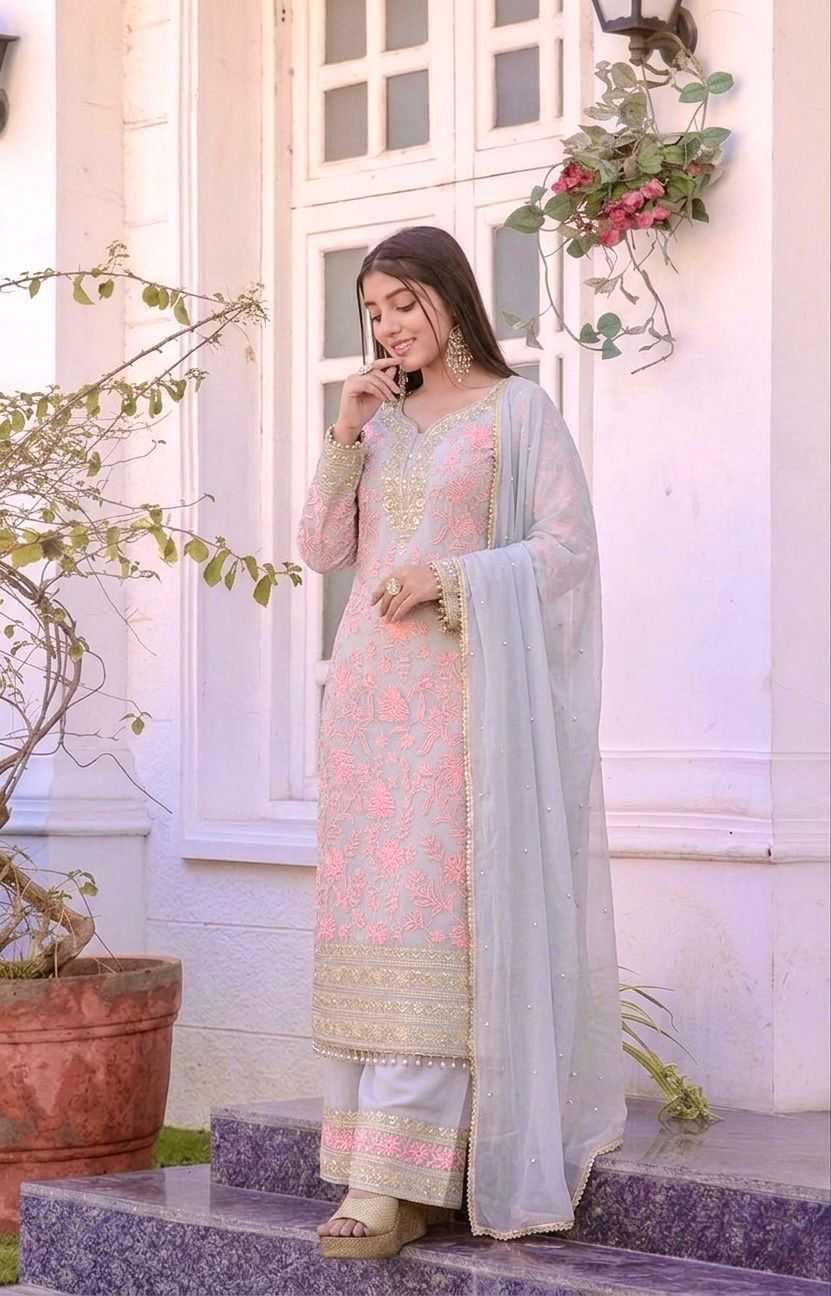 A woman stands on stone steps in front of a white building, wearing a light pink embroidered traditional outfit with a pale blue dupatta and beige flat shoes.