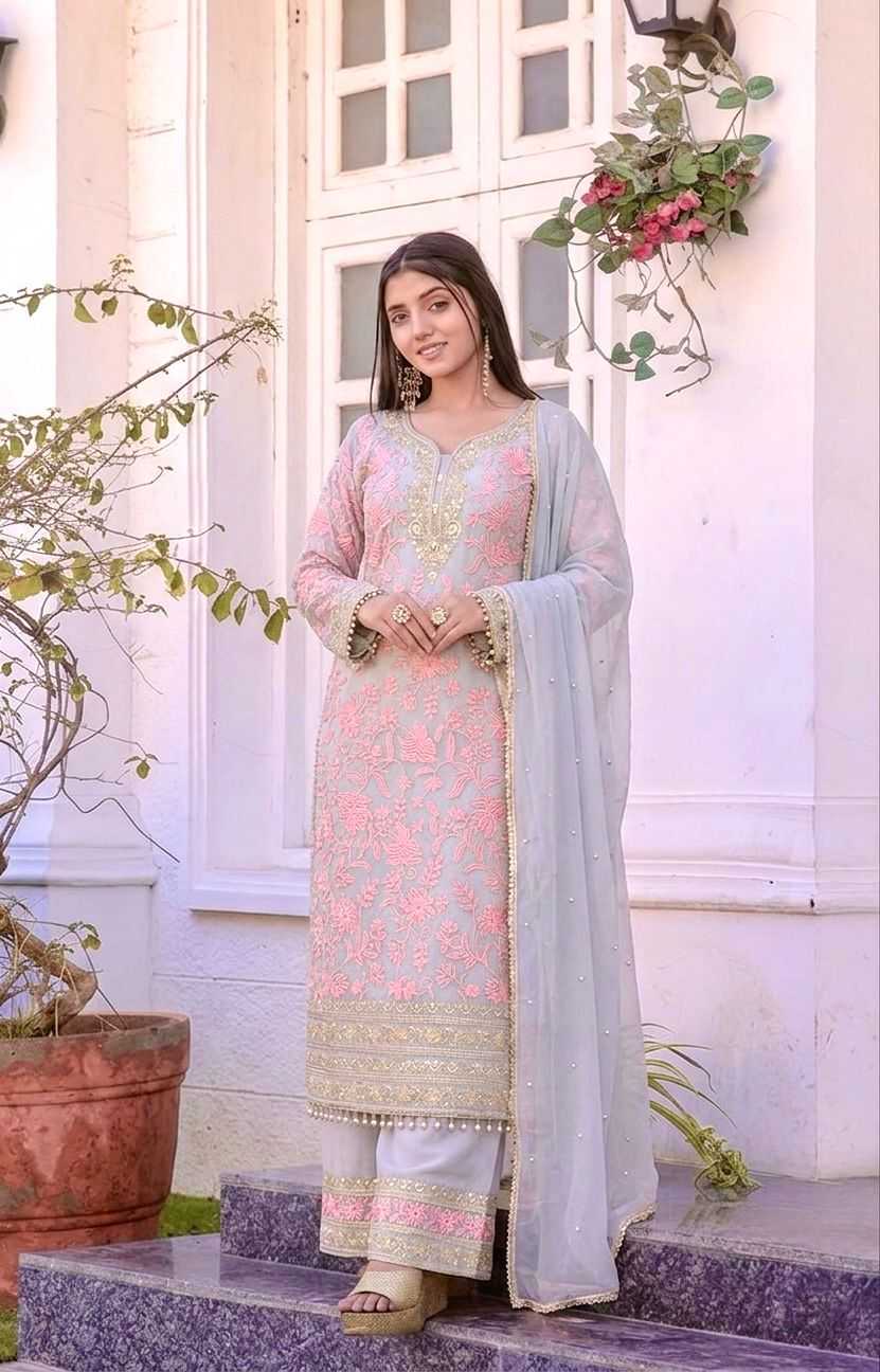 A woman stands on stone steps in front of a white building, wearing a light pink embroidered traditional outfit with a matching sheer light blue dupatta.