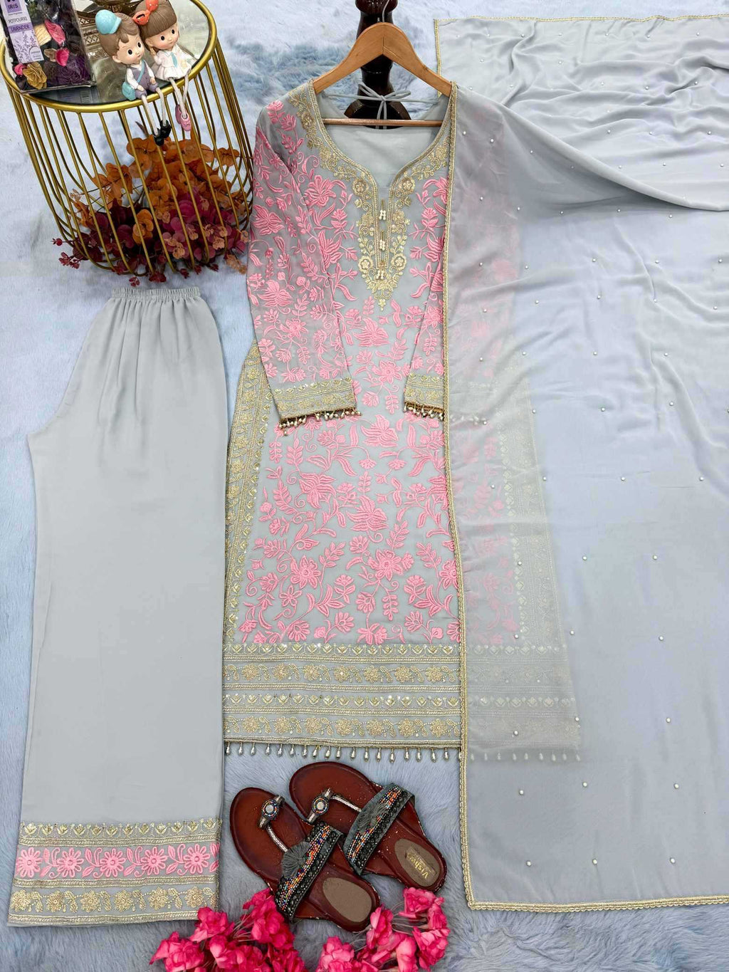 A three-piece women's salwar kameez suit set laid out on a light blue-gray surface. The set includes a light pink embroidered kameez top with gold zari detailing, light beige trousers with a pink and gold border, and a matching light gray/white dupatta.