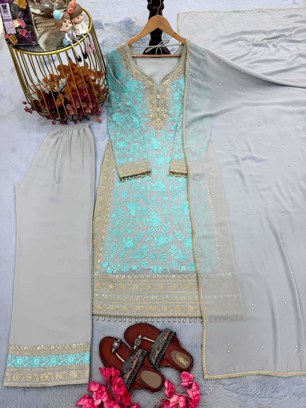 A traditional three-piece salwar kameez suit set laid out on a light gray surface. The set includes a mint green embroidered kurta with gold borders, matching beige trousers with gold and mint borders, and a sheer light gray dupatta.