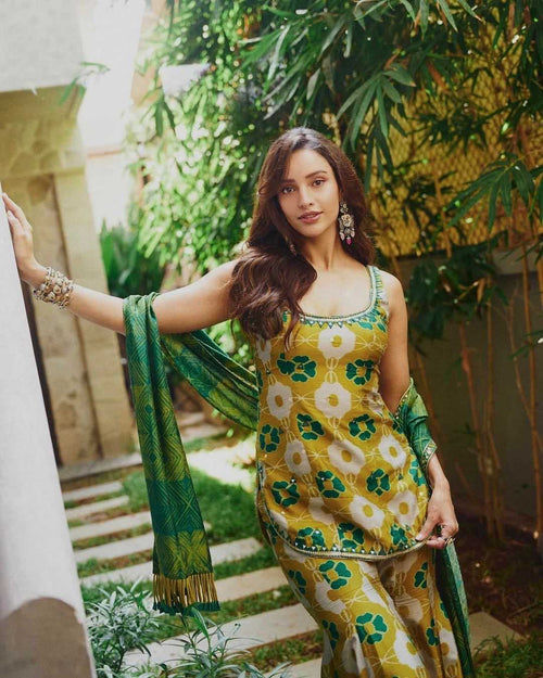 A woman models a yellow, green, and white printed traditional Indian salwar suit set with a matching dupatta in an outdoor garden setting.