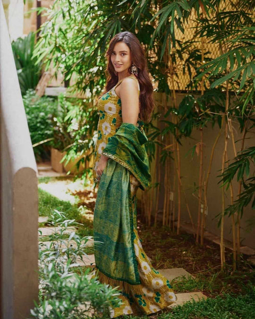 A woman stands in a garden courtyard wearing a green and yellow floral printed saree with a matching dupatta.