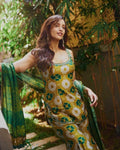 A woman wearing a yellow and green printed traditional outfit with a matching green dupatta, standing outdoors in front of bamboo and greenery.