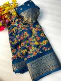 A folded blue saree with a vibrant floral and paisley print in yellow, red, and green, featuring a wide metallic gold border, displayed on a white fabric background next to yellow flowers.