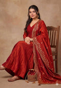 Woman in a red traditional outfit with gold embroidery sitting on a wooden chair against a beige background.