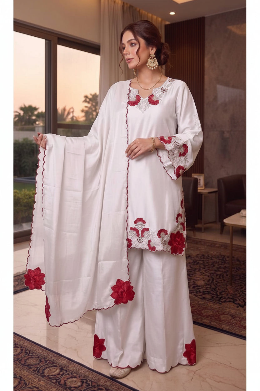 Woman wearing a white outfit with red floral embroidery, standing indoors.