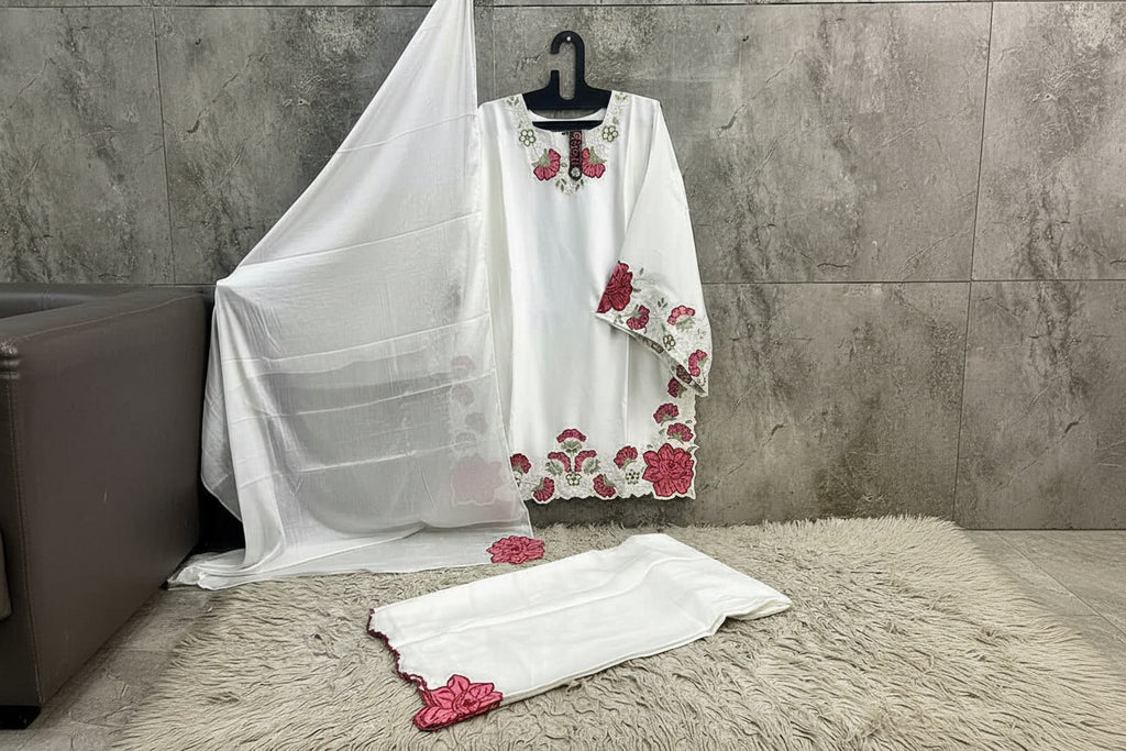 White embroidered dress with red floral patterns on a textured surface and gray wall background.