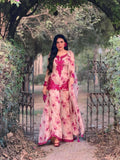 A woman stands in a garden archway wearing a traditional three-piece salwar kameez outfit in beige with vibrant pink floral embroidery and print, along with a matching dupatta.