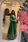 A woman wearing a green long-sleeved Anarkali gown with gold embroidery and a white collar, standing in a mirror within a boutique. Pink artificial flowers are visible on the right side of the frame.