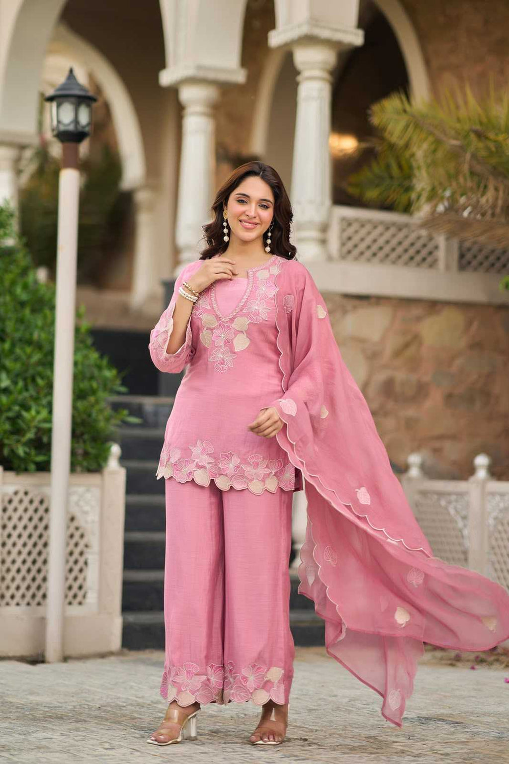 A woman modeling a pink traditional three-piece outfit with floral embroidery, featuring a kurta, wide-leg pants, and a matching sheer dupatta, posed outdoors in front of a building with arches and columns.
