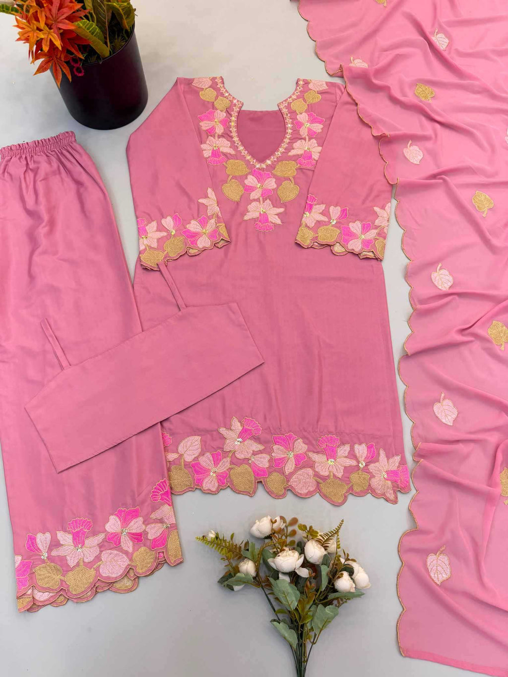 A pink traditional three-piece clothing set laid flat on a light gray surface, including a long kurta with floral embroidery, matching pants, and a draped dupatta. The embroidery features pink, yellow, and white floral patterns with scalloped edges.