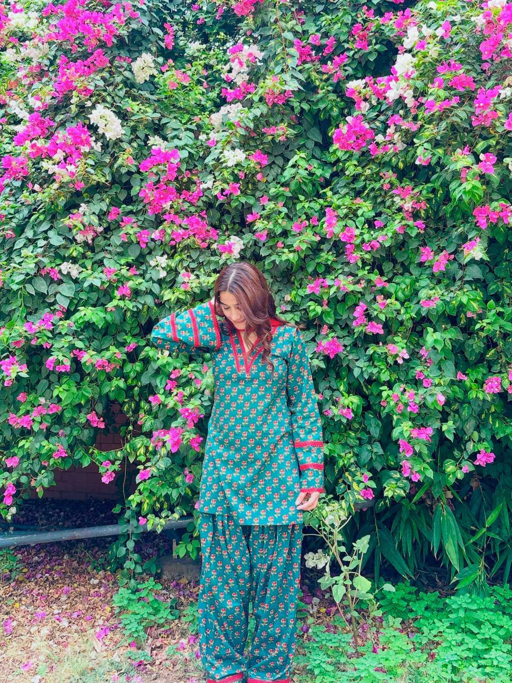 A woman wearing a green and white patterned kurta and matching pants stands in front of a dense wall of pink bougainvillea flowers.