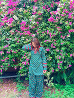 A woman wearing a green and white patterned kurta and matching pants stands in front of a dense wall of pink bougainvillea flowers.