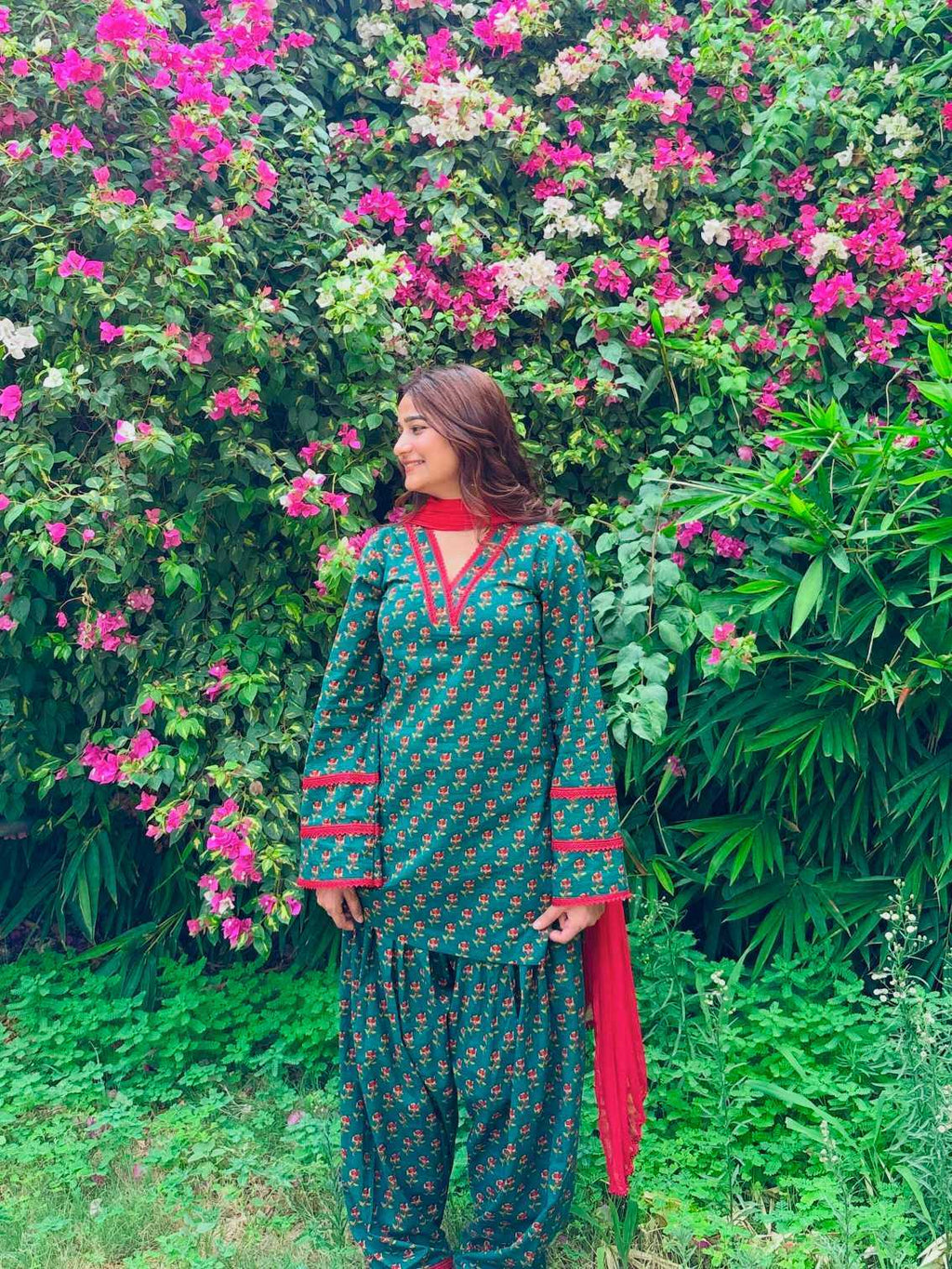 A woman wearing a green and red printed traditional outfit stands in front of a dense wall of pink flowers and green foliage.