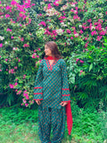 A woman wearing a green and red printed traditional outfit stands in front of a dense wall of pink flowers and green foliage.