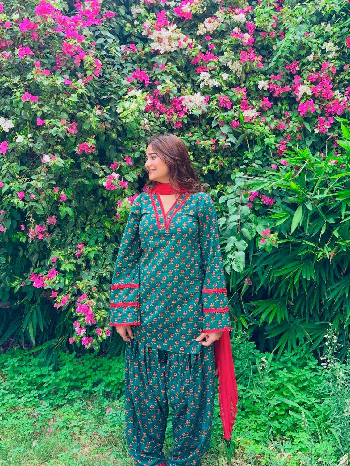 A woman wearing a green and red printed traditional outfit stands in front of a dense wall of pink flowers and green foliage.