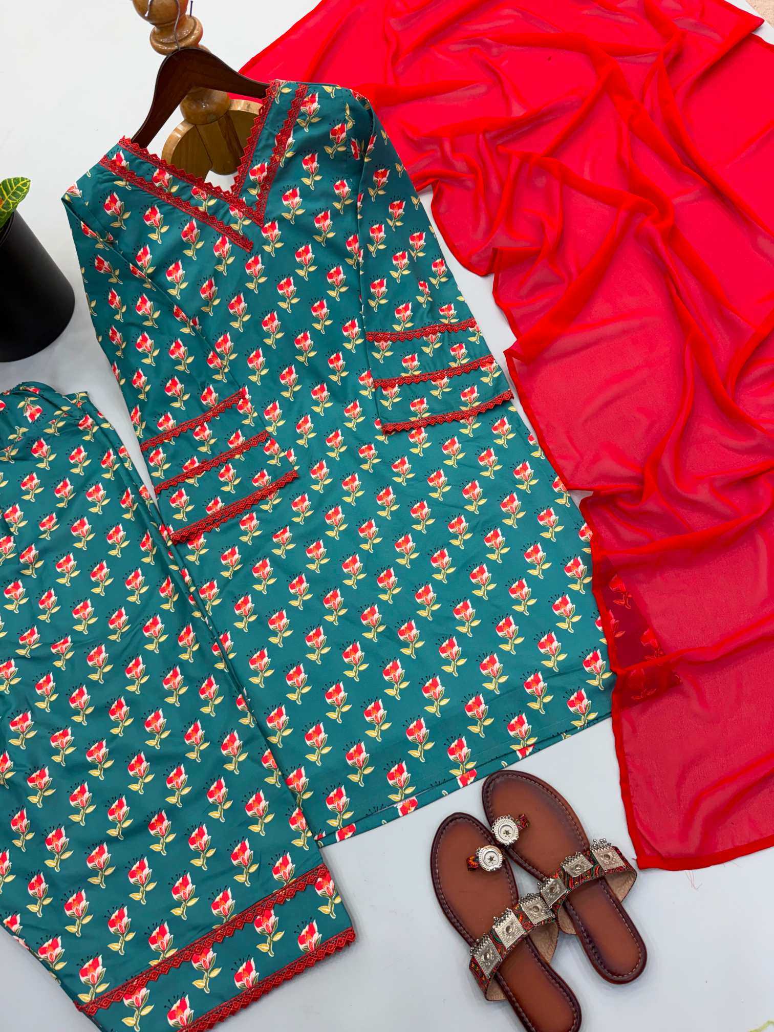 A flat-lay of a two-piece women's pajama set featuring a teal green base with a red and yellow floral print, styled with a bright red sheer scarf and brown leather slide sandals on a white background.