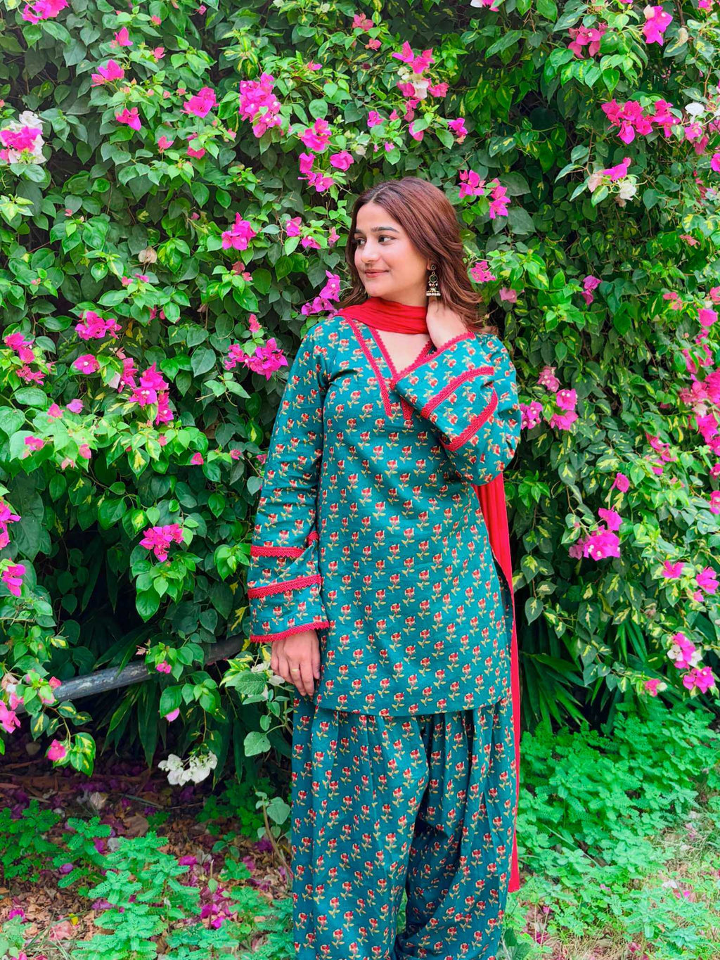 A woman wearing a teal and gold floral print kurta set with red trim, standing in front of a lush green garden wall covered in pink flowers.