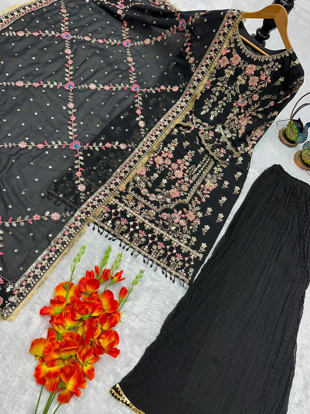 Black embroidered ethnic wear set featuring a kurta with floral embroidery, a matching dupatta with geometric beadwork, and black pants, arranged on a white textured surface with decorative flowers.