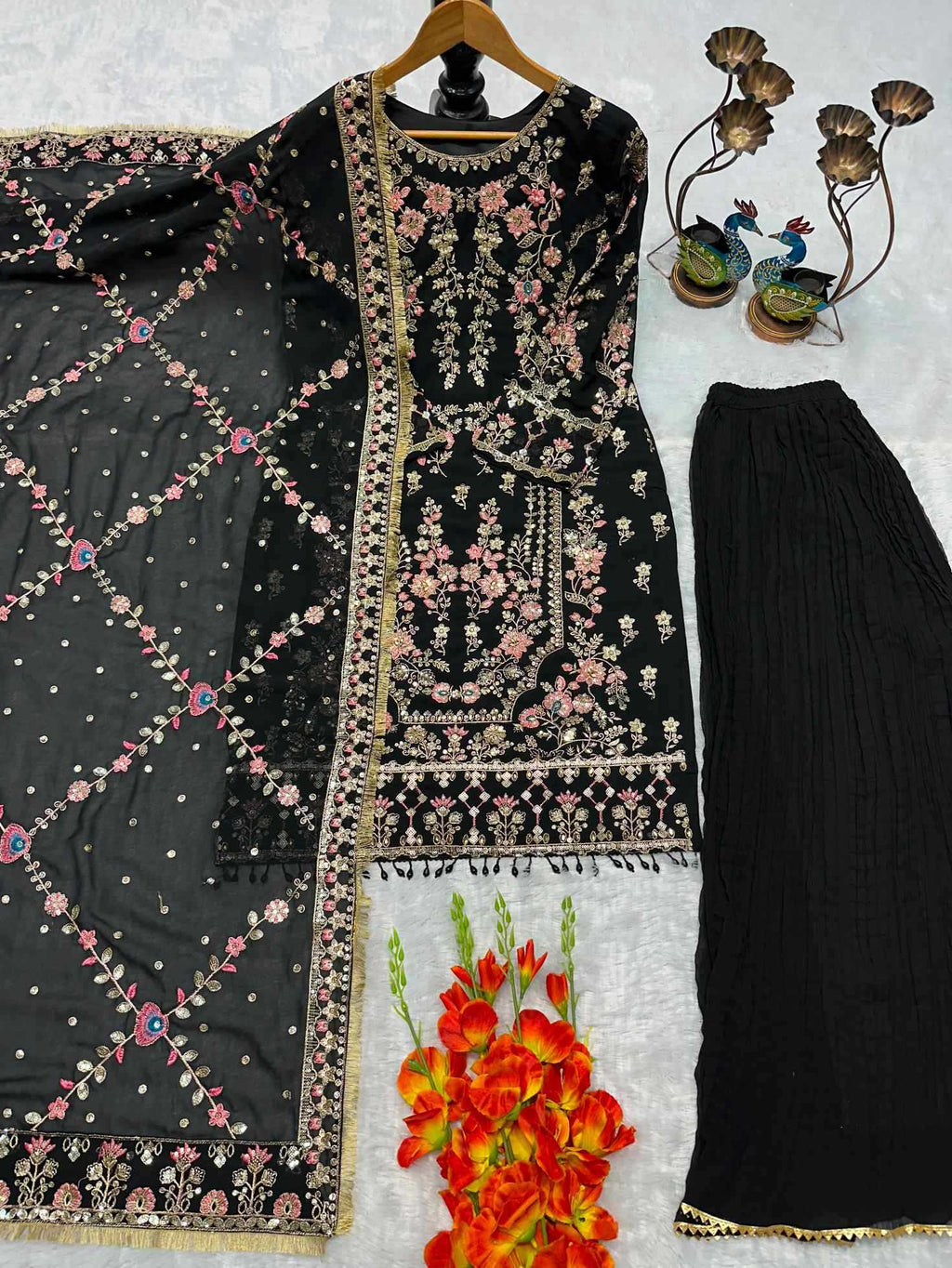 A black three-piece ethnic outfit laid out on a white surface, including a heavily embroidered kurta with floral and geometric detailing, a matching embroidered dupatta, and black pleated palazzo pants. Decorative flowers and a small peacock figurine are placed around the outfit.