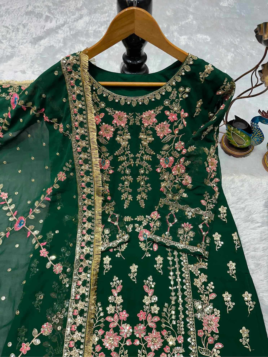 A dark green embroidered traditional dress with pink floral patterns and gold metallic detailing, displayed on a wooden hanger against a textured white background.