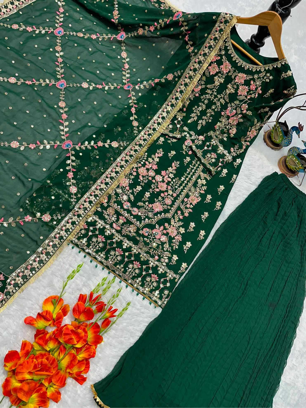 A dark green traditional embroidered outfit laid flat on a white surface, including a heavily embellished embroidered top, matching skirt, and sheer embroidered dupatta. The set is styled with orange flowers and small decorative items in the corners.