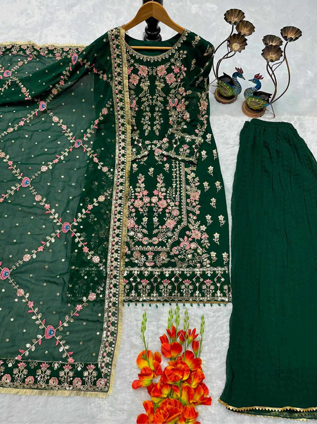 A dark green traditional three-piece outfit set featuring a heavily embroidered kurta with floral patterns, matching printed dupatta, and wide-leg trousers, arranged on a white textured background with decorative props.