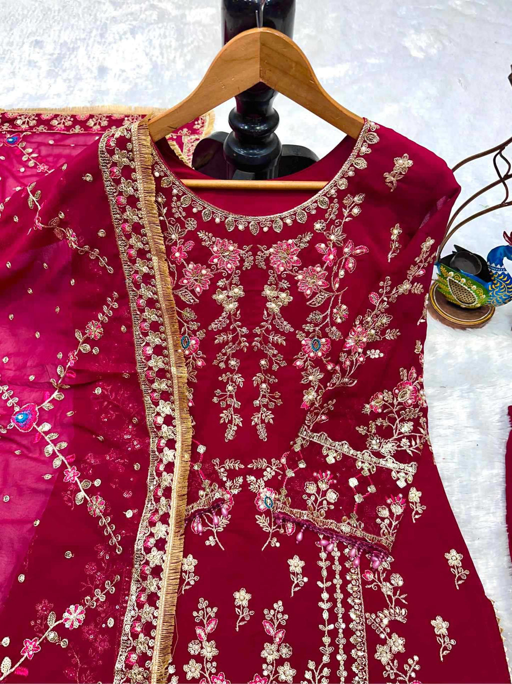 A deep magenta red embroidered kurta on a wooden hanger, featuring intricate gold floral embroidery, a gold border, and small blue and pink embellishments, set against a light textured background.