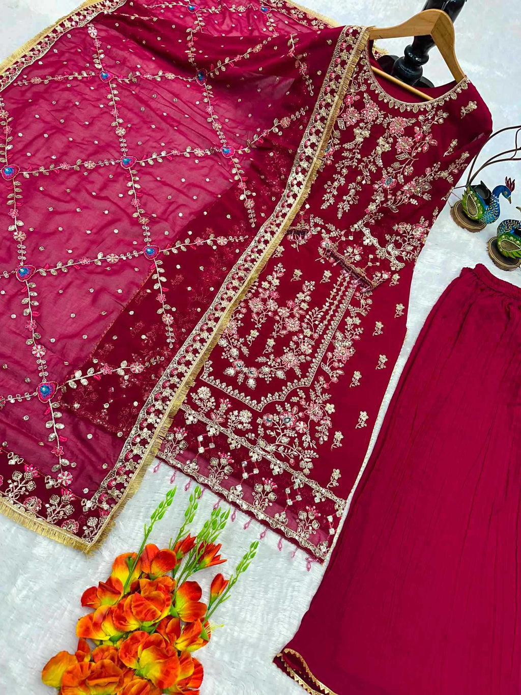 A traditional Indian salwar suit set in deep magenta pink, featuring an embroidered kurta with gold and silver mirror work, a matching dupatta with geometric patterns, and wide-leg pants. The set is styled on a white textured surface with decorative orange flowers and a pair of sunglasses.