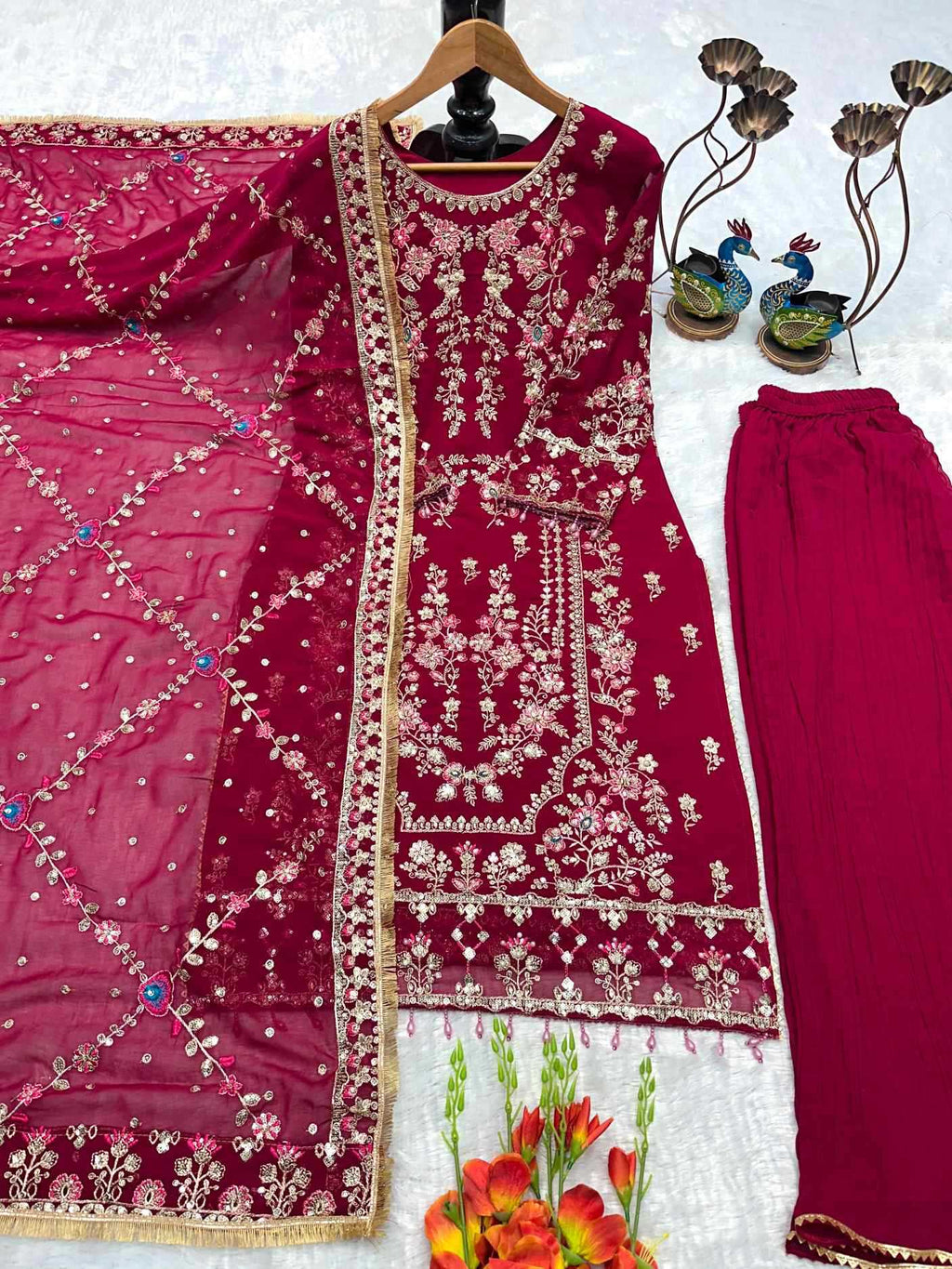 A deep magenta pink ethnic three-piece suit set featuring a heavily embroidered kurta on a hanger, matching trousers, and a sheer embroidered dupatta, arranged on a white textured background with decorative flowers and peacock figurines.