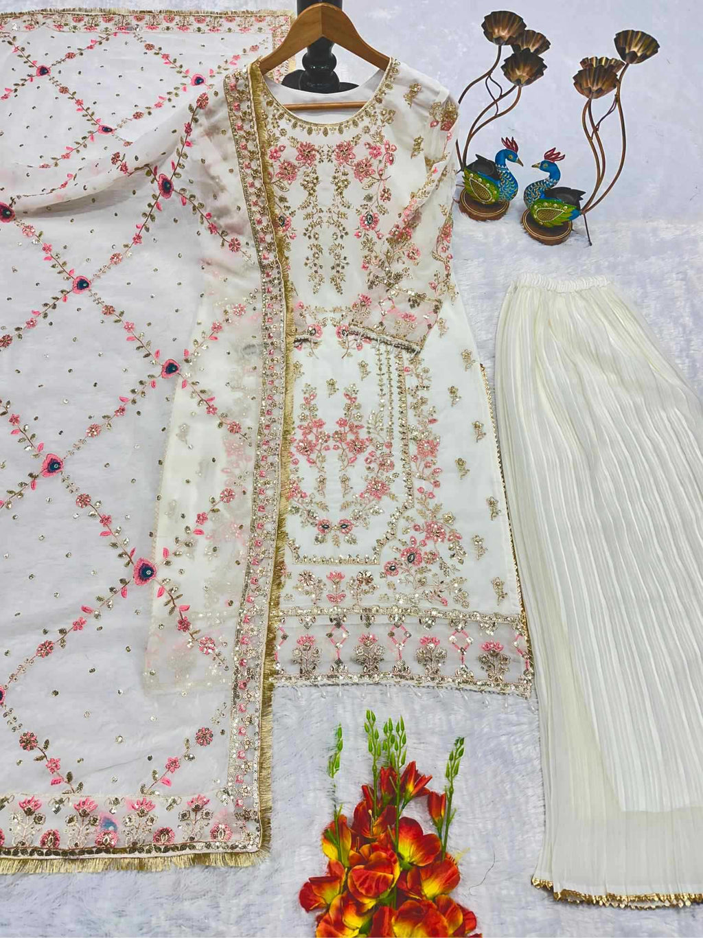 A three-piece women's ethnic wear set featuring an off-white embroidered kurta, matching pleated trousers, and a printed dupatta. The kurta is decorated with intricate pink and gold floral embroidery, paired with a sheer off-white dupatta featuring a pink and gold geometric lattice pattern.