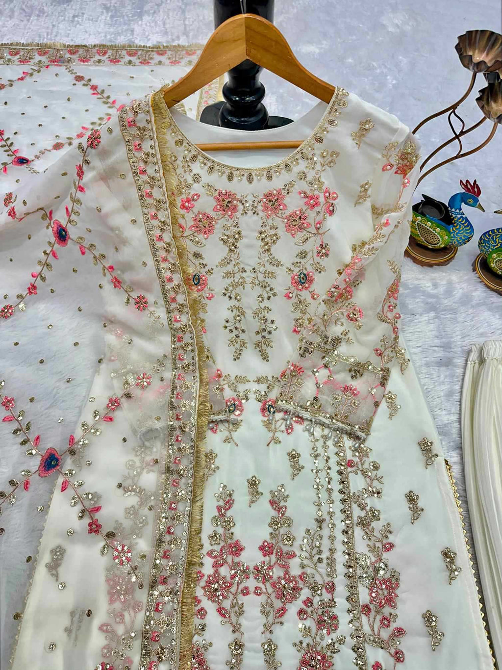 An off-white/cream traditional embroidered dress displayed on a wooden hanger, featuring intricate gold and pink floral embroidery with sequins and mirror work, accompanied by a matching sheer dupatta.