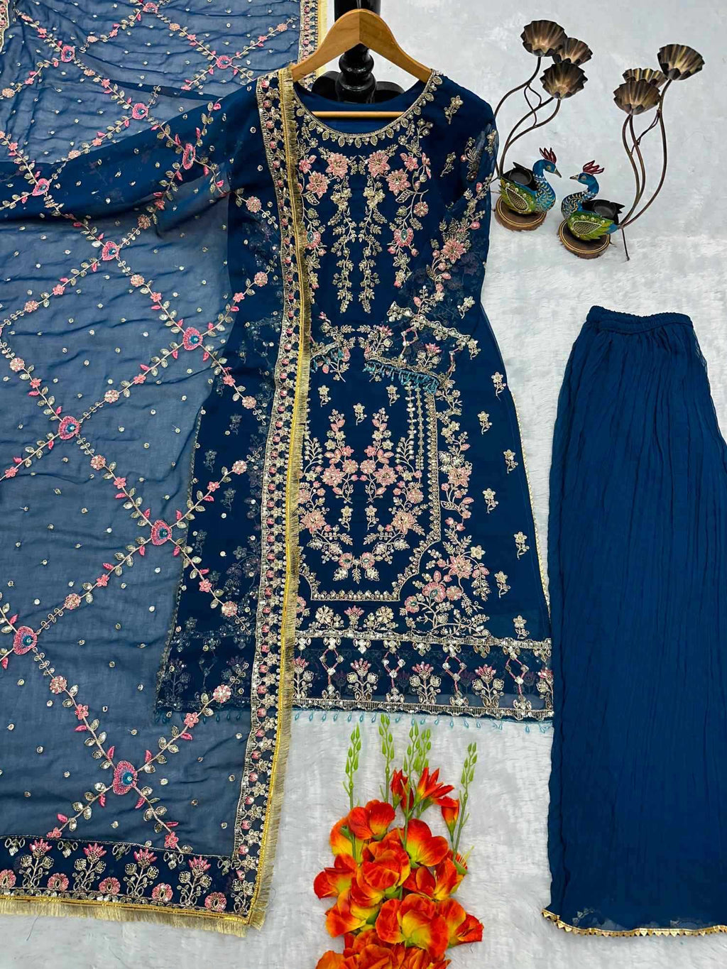 A navy blue three-piece salwar kameez suit set with intricate floral embroidery and gold borders, displayed on a white textured background with decorative flowers and peacock figurines.