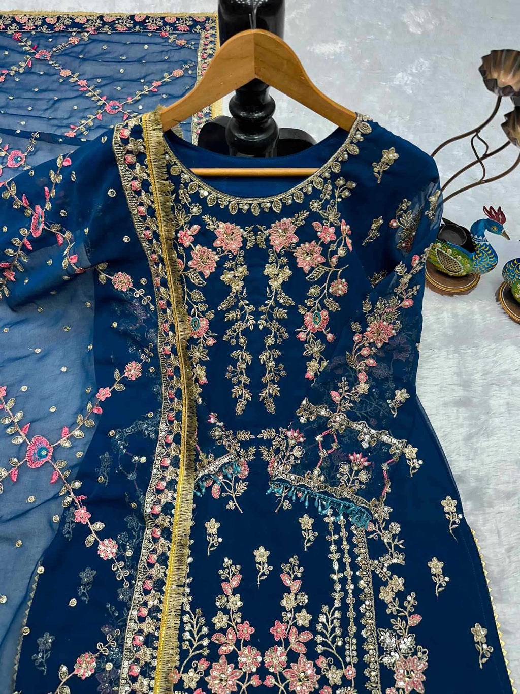 A dark blue embroidered kurta with pink floral patterns and gold zari detailing, displayed on a wooden hanger against a light gray background.
