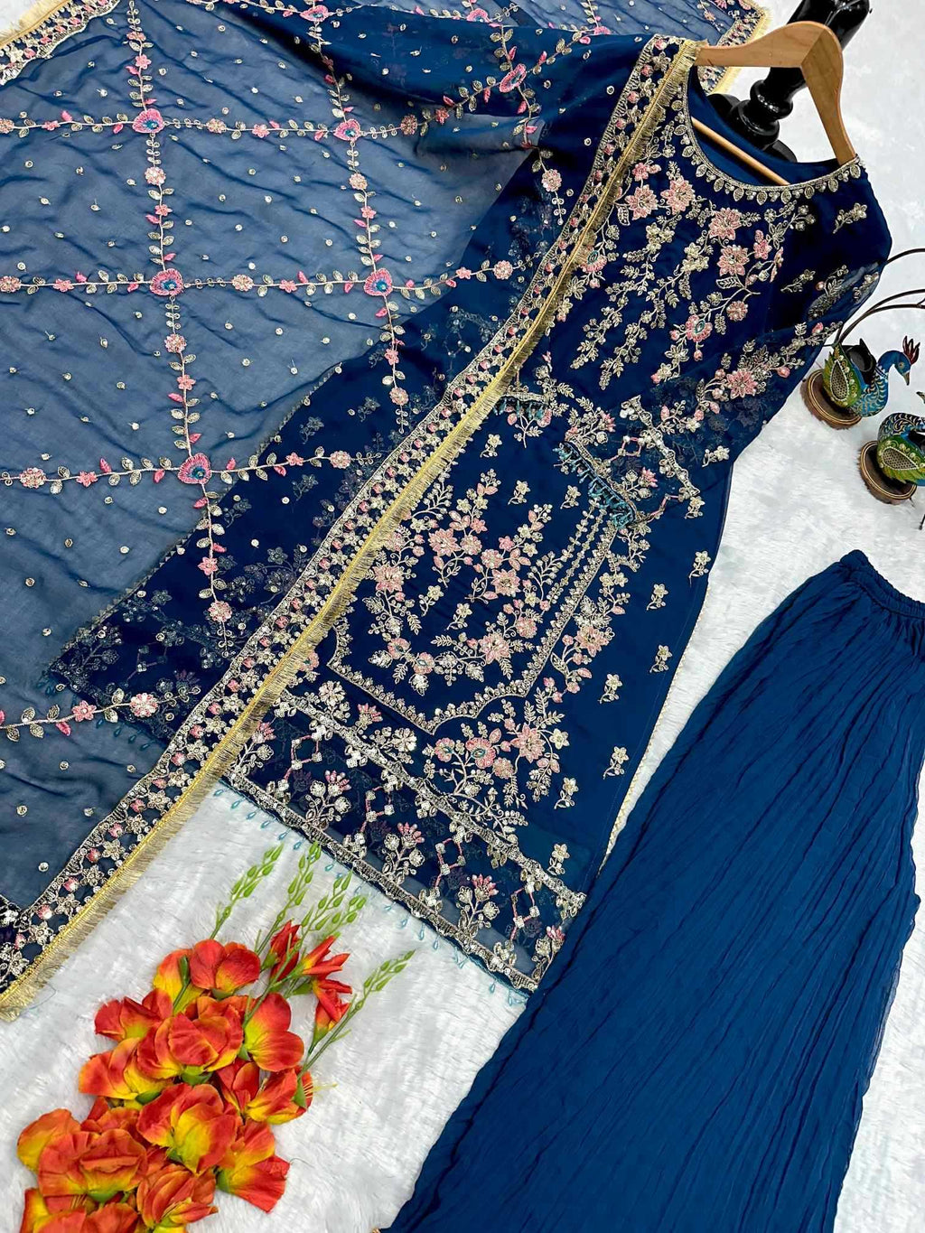 A navy blue ethnic suit set laid out on a white textured surface, including an embroidered kurta with floral detailing, matching pleated pants, and a sheer embroidered dupatta. Decorative flowers and small accessories are placed around the outfit.